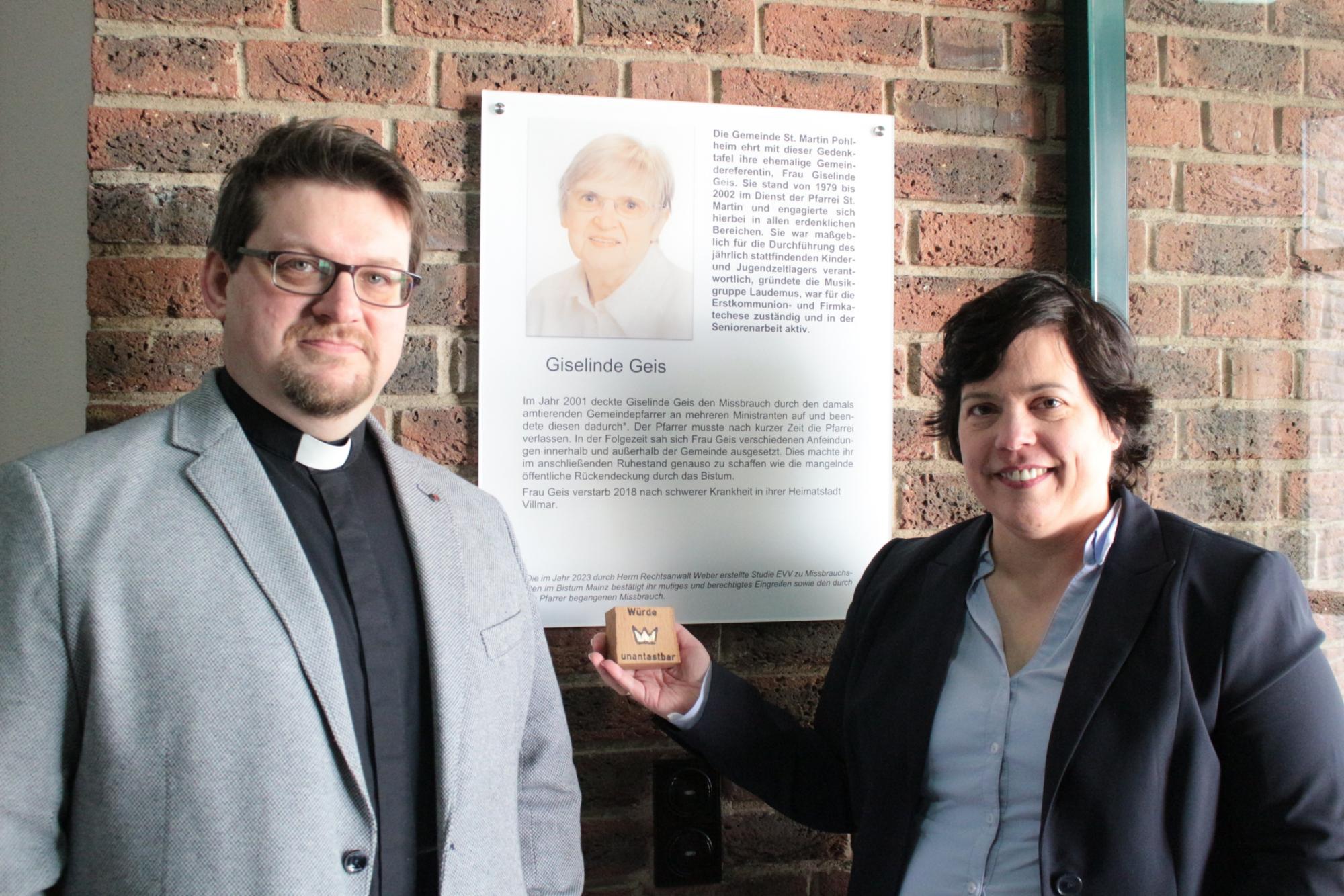 Pohlheim, 25. Februar 2024: Pfarrer Martin Sahm (links) und die Bevollmächtigte Stephanie Rieth vor der Gedenktafel für Giselinde Geis im Pfarrzentrum von St. Martin. (c) Bistum Mainz / Blum