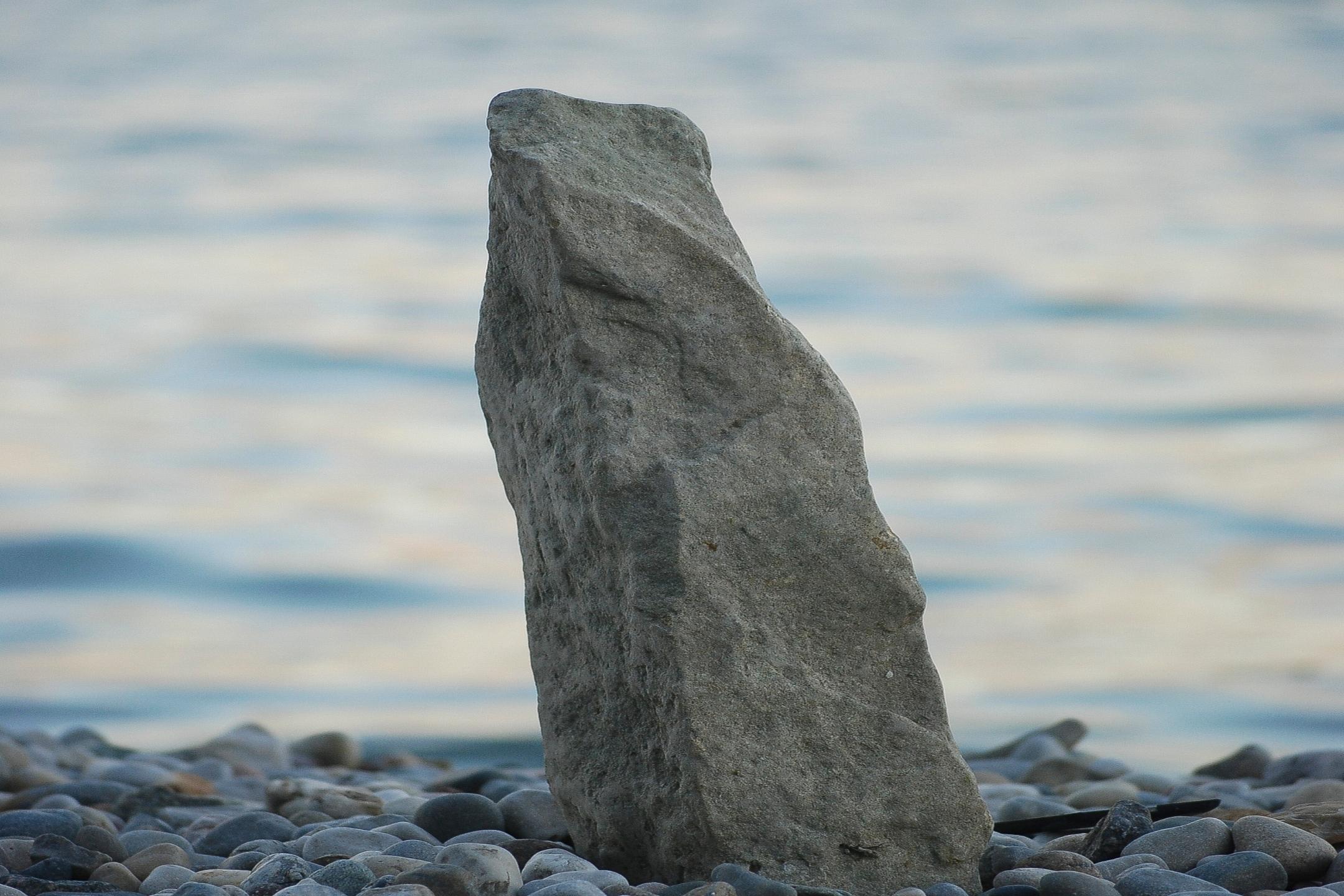 Ein größerer, aufragender Stein steht im Kiesstrand am Ufer eines Meeres. Das Bild strahlt Ruhe, Standfestigkeit und Unerschütterlichkeit aus.